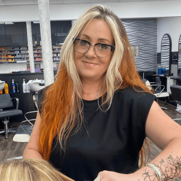 Kerry, Advanced Senior Stylist at Hair Pro UK in Bexhill-on-Sea, smiling warmly in front of the salon’s branded backdrop, representing decades of colour mastery, professional expertise, and long-standing dedication.